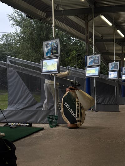 The image shows a golf driving range in South Ribble, United Kingdom (coordinates: 53.771, -2.57). A person in light-colored pants is partially obscured by a large TaylorMade golf bag in the foreground, standing near a covered hitting bay with a gray mesh net and a series of golf balls laid out on the green carpet floor. The person is facing toward the hitting bay and appears to be swinging a golf club. Several electronic scoreboards displaying golf footage are mounted on poles above the hitting bays. The scene is located under an open, metal-framed structure with overhead lights and a gray concrete floor. The weather is overcast and possibly raining. The location is a golf driving range.