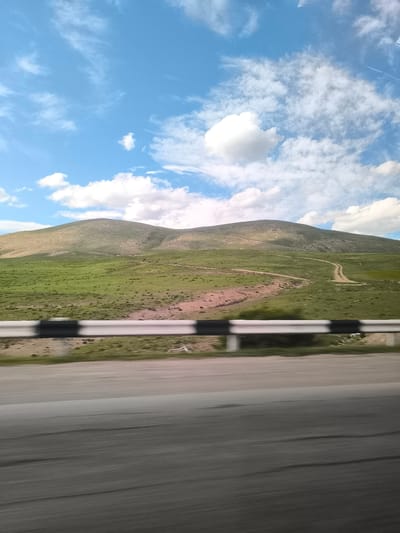 This image captures a vibrant, expansive landscape as seen from a moving vehicle in Yelpin, Armenia. The scene is dominated by undulating, green hills under a bright blue sky dotted with scattered white clouds, some appearing wispy while others are more defined cumulus.
In the foreground, a heavily motion-blurred asphalt road indicates travel, and a black and white striped metal guardrail runs across the mid-ground, also exhibiting motion blur. Beyond the guardrail, the rolling hills rise, covered primarily in lush green grass, with some patches of sparser, drier-looking vegetation. Several unpaved dirt tracks or paths can be seen winding through the verdant terrain of the hills, suggesting routes for off-road vehicles or hikers.
The time of day appears to be late morning or early afternoon, characterized by bright, even daylight. The weather is clear and sunny with pleasant cloud cover. The overall impression is one of a vast, natural, and relatively unpopulated mountainous region. There are no people or discernible text visible in the image. The image conveys a sense of a journey through scenic, rugged natural beauty.