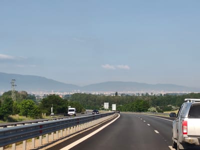 The image shows a highway leading towards a city with mountains in the background. The highway has multiple lanes and is bordered by a metal guardrail. Several vehicles are visible on the road, including a white van, a black car, and a silver pickup truck. The surrounding landscape is lush with green trees and vegetation. In the distance, a city skyline is visible with buildings and a prominent power line tower. The sky is clear and blue, with a few scattered clouds. The image appears to be taken from the perspective of a moving vehicle. The specific location is the highway leading to Dolni Bogrov, Bulgaria.