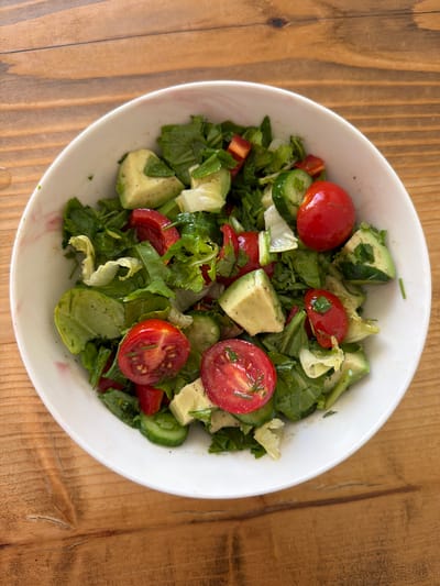 A close-up, overhead shot captures a white bowl filled with a vibrant salad. The salad is composed of mixed greens, diced avocado, sliced cherry tomatoes, and cucumber. Diced red bell pepper pieces are also visible. The ingredients are lightly tossed, with visible flecks of seasoning, possibly salt and pepper. The bowl itself has a subtle, marbled pink rim. It rests on a rustic wooden surface with visible grain and a few subtle cracks. The lighting suggests an indoor setting, likely a well-lit room. There are no people or animals present in the image. The scene is static and focused on the food. This image was taken in Zeytinburnu, Turkey.