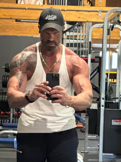 The image is a selfie of a muscular man taken in a gym in Jemelle, Belgium. The man is standing upright, looking directly into his dark-colored smartphone, which he holds in both hands to capture his reflection. He wears a dark baseball cap, a plain white tank top, and dark athletic pants or shorts.
His physique is prominently featured, showcasing well-developed muscles in his arms and shoulders, with visible veins. Intricate, dark-patterned tattoos adorn his left arm. He accessorizes with a thick gold chain necklace and a gold bracelet on his right wrist, along with a dark watch on his left wrist. His facial hair includes a beard, and his expression is neutral and focused.
The background of the image reveals a typical gym environment, including various metal frames of workout equipment and stacked weights. The lighting is bright and even, characteristic of an indoor setting, possibly with natural light filtering in.
Visible text on his baseball cap is mirrored, appearing as "ƆIЯⱭ DЯO⅃", which would read "LORD DRICH" or "LORD RICH" when un-mirrored. A small red and white logo, resembling the Champion brand, is discernible on his dark pants. The image captures a moment of self-documentation, likely during or after a workout.