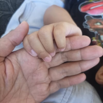This image displays a close-up view of an adult's hand and an infant's hand in contact. The reported location is Pasir Muncang, Indonesia.
An adult hand, exhibiting a light brown skin tone with visible creases and knuckles, is positioned at the lower-left and center of the frame, with its palm facing generally upwards and its fingers extending towards the upper-right. An infant's smaller, plump hand, also with a light brown skin tone, is positioned above the adult's hand. The infant's fingers are curled around and gripping the adult's index finger. Short, unpainted fingernails are visible on both the infant's and adult's digits.
The background consists of various indistinct fabric elements. In the upper-left, a dark, textured fabric is partially visible. Towards the upper center, a section of light blue fabric, consistent with an article of clothing, is present. On the right side, a dark fabric with indistinct, multi-colored graphic patterns, resembling a design on a t-shirt, is visible. The lower-right and central portions of the background feature a light grey or khaki-colored fabric, possibly trousers, with visible stitching.