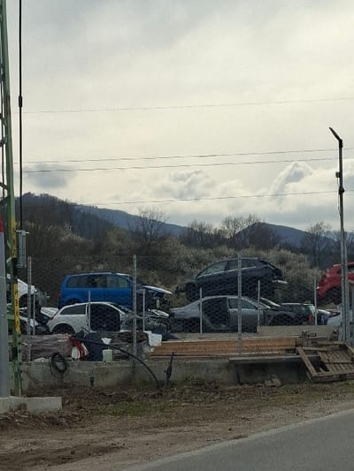 This image depicts an outdoor automobile salvage yard, also known as a junkyard, located in Cherniche, Bulgaria. The scene is presented under a heavily overcast sky, with mountains visible in the distant background.

In the immediate foreground on the left, a section of a green metal lattice structure, likely a utility pole or support, is partially visible. On the right, a black utility pole with a street light fixture and an attached solar panel stands vertically.

A chain-link fence, erected on a concrete foundation, separates the foreground area, which features a dirt patch and an asphalt road on the far bottom right, from the salvage yard. Within the fenced area, numerous damaged and partially dismantled vehicles are stacked and scattered. Prominently visible vehicles include a blue SUV, a white hatchback, a dark grey sedan with its front end removed, and a dark blue/black sedan stacked atop other vehicles. A red vehicle is partially visible on the far right. Below the fence line, on the concrete base, discarded items such as a black hose or cable, red and white fabric, and wooden planks and a pallet are present.

The background features bare deciduous trees and shrubs covering the lower slopes of mountains that extend across the horizon. No individuals are visible within the scene, and no active interactions or movements are depicted. The overall environment suggests a rural or semi-rural setting.