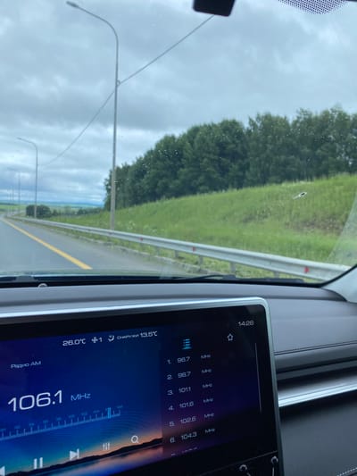 The image is taken from inside a car, looking out at a cloudy, overcast sky and a rural landscape. A highway with a yellow line is visible, bordered by a guardrail and green grassy hills with trees in the background. The car's dashboard features a large infotainment screen displaying the current radio station as "106.1 MHz" and a list of other frequencies. The screen also shows the time as "14:28", the outdoor temperature as "13.5°C", and the indoor temperature as "26.0°C". There are water droplets on the windshield, suggesting it may be raining or has recently rained. The location is identified as Александровское, Russia. There are no people or animals visible in the image.