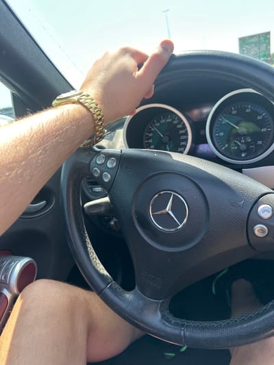 The image shows the interior of a Mercedes-Benz car. The dashboard is visible, with the steering wheel featuring the Mercedes-Benz logo prominently displayed. The speedometer and fuel gauge are partially visible behind the steering wheel. A male hand with a gold watch is gripping the left side of the steering wheel. Part of the man's legs are also visible. There is no information to determine that this image was taken in Leidschendam, Netherlands.
