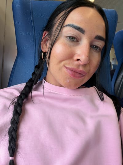 In the city of Ушаки, Russia, a young woman with dark hair, styled in two braids, is captured in a close-up selfie. She is seated on a blue textured chair, likely in a public transport setting, given the visible headrest and seat fabric. The woman is wearing a pink long-sleeved shirt. Her facial features are the focal point, with her eyes, eyebrows, and lips clearly visible. She is wearing gold hoop earrings. A small blemish is noticeable on her chin. The lighting suggests an indoor environment, possibly during the day.