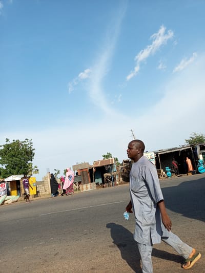 A man in a light gray traditional outfit walks across a paved road in Gadao, Nigeria. He holds a small, clear plastic bottle of water in his right hand. The man is bald and is walking towards the right of the frame. He is casting a long shadow on the road, indicating the sun is low in the sky, likely late afternoon or early morning. The weather appears clear with a bright blue sky dotted with a few wispy clouds.

In the background, several small shops or stalls line the side of the road. Some have corrugated metal roofs and open fronts, displaying various goods. A few people are visible near these stalls. To the left, a young man in a striped t-shirt and shorts stands near a yellow structure with signs. Further down the road, a woman in a pink headscarf and a patterned dress is walking with another woman in a black hijab.

Behind the stalls, there are some trees and vegetation. Power lines are also visible stretching across the scene. The overall setting suggests a market or roadside commercial area in a Nigerian town. The mood appears calm and ordinary, depicting daily life. Some text is visible on signs, including "POS CENTER" on the yellow structure.