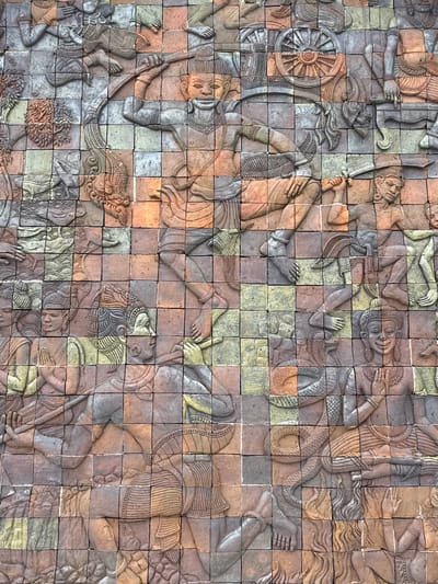 The image displays a detailed bas-relief mural made of terracotta tiles, depicting a scene with multiple figures engaged in various activities. The mural is located on a wall in Chang Khoeng, Thailand.
In the upper left quadrant, a figure is shown in a dynamic pose, possibly in motion, with their arms outstretched and holding a weapon. To the right of this figure, another individual is depicted with their hands raised, holding what appears to be a bow. A large wheel, reminiscent of a chariot wheel, is visible in the upper right portion of the mural.
The central area of the mural features several figures in different stances. One figure is shown playing a flute, while another appears to be wrestling or engaged in a struggle with a serpentine creature. Several other figures are depicted with their hands in prayer or offering gestures.
The lower portion of the mural continues the narrative with more figures. A figure is shown with a bow and arrow, aiming at something unseen. Another figure is depicted in a reclining or resting position, with hands clasped. The serpentine creature from the central area continues to be depicted, coiling around some of the figures. The mural is composed of individual square tiles, each contributing to the overall composition and narrative. The color palette is dominated by earthy tones of terracotta, with some areas exhibiting hints of green and grey, likely due to weathering or the original coloring of the tiles. The lighting suggests an outdoor environment, with shadows indicating the direction of light.