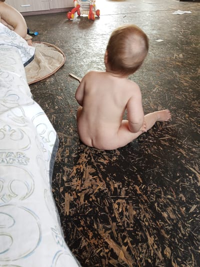 A high-angle, full shot captures a baby in a diaper seated on a dark, patterned floor, looking away from the camera. The baby's back is to the viewer, and the head is turned slightly to the right. The baby has light brown hair. The floor appears to be made of dark wood chips or a similar textured material.

To the left, a portion of a white bedsheet with light blue and grey patterns is visible, including a section with the text "STAR WARS" in stylized block letters. In the background, further to the left and slightly behind the bedsheet, another person or baby is partially visible, also seated or lying down.

In the upper background, part of a light-colored furniture piece with two cabinet doors can be seen. To the right of this furniture, a red and yellow baby walker is partially visible. A small piece of white paper or tissue is on the floor in the upper right quadrant of the image. The lighting suggests an indoor setting, likely during the day. The scene appears to be a casual, everyday moment in a home. The focus is on the baby in the foreground, with other elements providing context to the domestic environment.