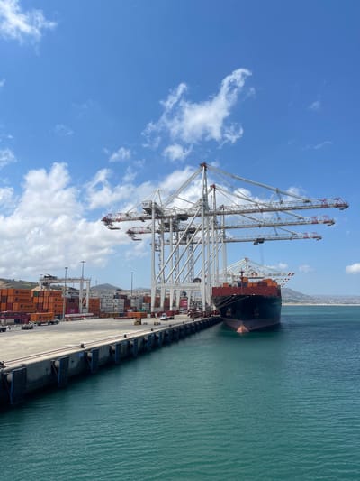 The image depicts a bustling container port under a bright, partly cloudy sky. In the foreground, the teal-colored water of the harbor laps against a concrete pier. The pier is lined with large, dark blue fenders attached by chains, with some bearing the marking "B723". Along the edge of the pier, there are various shipping containers stacked high, predominantly orange and white, with visible branding such as "Hapag-Lloyd" and "ONE". Several trucks and other port vehicles are visible amongst the containers.
Dominating the scene are massive, white gantry cranes, also known as port cranes, with their long, articulated arms extended over the pier and water. One of these cranes is actively positioned over a large cargo ship docked at the pier. The ship, painted in dark red and black, is laden with more orange shipping containers stacked on its deck. The hull of the ship is visible, as is its bow, pointed towards the right of the frame.
In the background, beyond the immediate port area, a hilly, green landscape meets the horizon, suggesting the proximity of land to the port. The overall impression is one of industrial activity and global trade taking place at a modern maritime facility. The location is identified as Ksar al Majaz, Morocco.