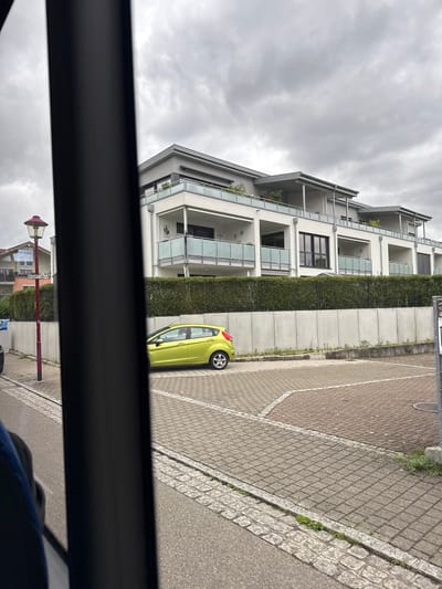 In the city of Bad Säckingen, Germany, a bright lime green Ford Fiesta is parked in a paved lot. The car is facing right and its front wheel is turned slightly towards the right. The building behind the car is a modern multi-story structure with balconies and large windows. A dense green hedge runs along the front of the building, separating it from the parking area. To the left of the car, a red lamppost stands on a sidewalk. The sky is overcast with gray clouds.