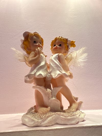 The media file displays a close-up, rotated view of a decorative figurine depicting two cherubs, likely made of resin or ceramic, against a textured, pale pink background. The cherubs are styled as infants with blonde hair and rosy cheeks, adorned with small white feathers suggesting wings. They are depicted in a playful, entangled pose, with one cherub resting on the other. Both are dressed in flowing white garments that appear to be made of a fabric-like material, possibly with glitter accents on the lower figure's base. The lighting appears to be artificial, creating soft shadows and highlighting the glossy finish of the figurines. The overall impression is one of innocence and childlike charm. No visible text is present in the image. The location context is Lopaskyne, Ukraine, but this information is not visually inferable from the image itself.
