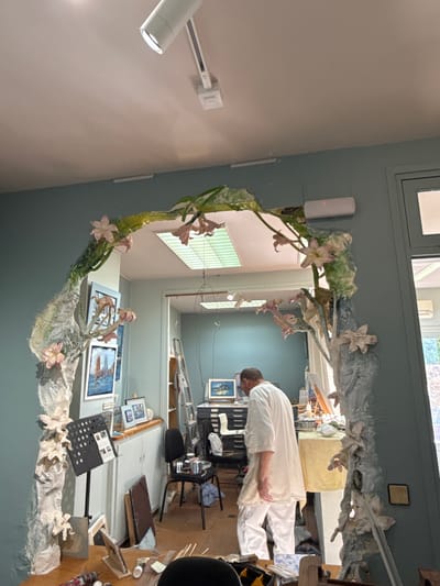 The image depicts an indoor scene, likely an artist's studio, with a person dressed in white working in the background. The foreground is dominated by a large, sculptural archway covered in a leafy, floral motif. The archway appears to be made of plaster or a similar material and is adorned with pink and white flowers.
The studio space behind the archway contains various art supplies and equipment. Paintings are visible on the wall, and a ladder leans against a shelf unit. The overall environment suggests a creative workspace filled with ongoing projects. The person in the background appears to be engaged in some activity related to the studio's purpose, possibly working on or near the archway. The lighting suggests a daytime setting, with overhead lights and natural light filtering in from unseen windows.
There is no information in the image that places the scene in Tossa de Mar, Spain.