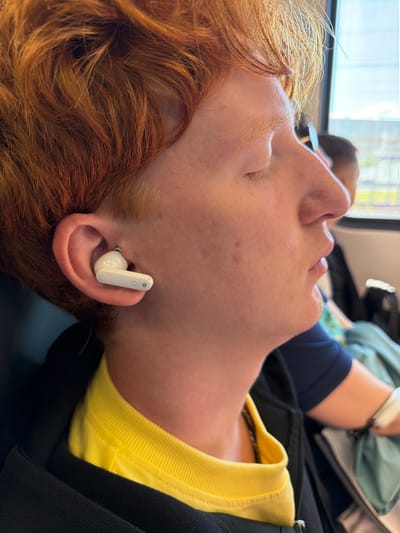 The media file is a close-up, eye-level shot of a person with reddish-blonde hair wearing a white earbud in their right ear. The person is lying down, with their head tilted back, exposing their profile. Their eyes are closed, and their facial expression appears relaxed. The earbud is white and has a stem. The person is wearing a bright yellow t-shirt, visible around their neck and shoulder, and a black jacket or sweater is layered over it. The background is blurred, but suggests an indoor setting with some light coming from a window, possibly indicating daytime. There is no visible text in the image. The context provided is "Bittsa, Russia," which offers a geographical location but no specific environmental cues visible within the image itself. The overall mood is peaceful and still, suggesting a moment of rest or listening.