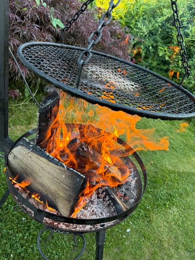 A close-up, eye-level shot captures an outdoor fire pit with a grilling grate suspended above it, located in Mordarka, Poland. The grate, made of black metal mesh, is tilted, revealing the glowing embers and vibrant orange flames of the burning logs below. Several pieces of firewood are visible within the fire pit, some charred and breaking apart. The background consists of lush green grass, with foliage in shades of red and green framing the scene. The overall impression is one of a rustic outdoor setting with a fire actively burning.