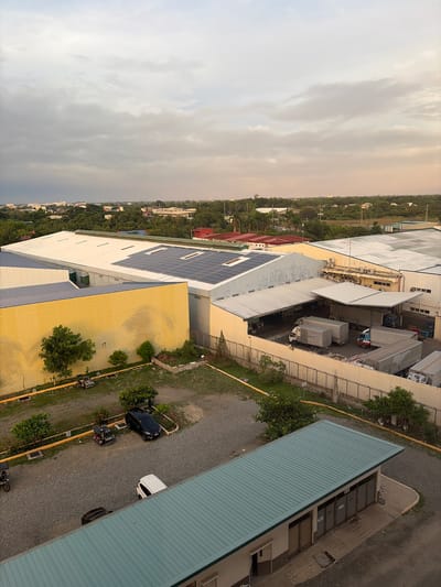 This is an aerial, oblique view of an industrial area in Dagupan, Philippines, under a cloudy sky with hints of sunset colors. The scene is dominated by large, multi-colored warehouse-style buildings with corrugated metal roofs. One prominent building has a bright yellow facade, while another, larger structure features a white roof covered in solar panels. Several trucks, including box trucks and refrigerated units, are parked at loading docks. In the foreground, a smaller building with a teal-colored metal roof and what appear to be multiple garage doors is visible. The ground is covered in gravel, and there are patches of grass and scattered trees. Several vehicles are parked in a lot, including a black SUV, a white van, and a couple of motorized tricycles. The overall impression is one of a functional industrial or commercial zone.