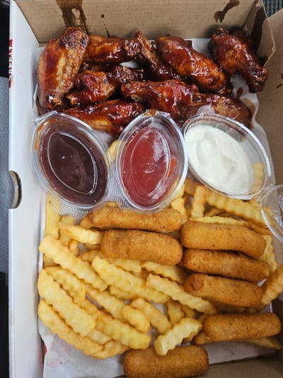 A bird's-eye view shows a takeout box filled with various fried foods. The box contains chicken wings coated in a dark, glossy sauce, golden-brown french fries with a crinkle cut, and several breaded, finger-shaped fried items that appear to be mozzarella sticks or chicken tenders. Three small, clear plastic containers are nestled among the food. One holds a white dipping sauce, likely ranch or mayonnaise. Another contains a bright red sauce, probably ketchup, and the third holds a dark brown sauce, possibly barbecue sauce. The food is arranged in a casual, haphazard manner, typical of a takeout order. The background is the inside of a cardboard box, with some white paper liner underneath the food. The lighting suggests the photo was taken indoors or under an overcast sky, as there are no strong shadows. The scene conveys a sense of casual dining and comfort food. There is no visible text in the image. The location context provided is Gornji Kokoti, Montenegro, but there are no visual cues in the image itself to confirm this location.