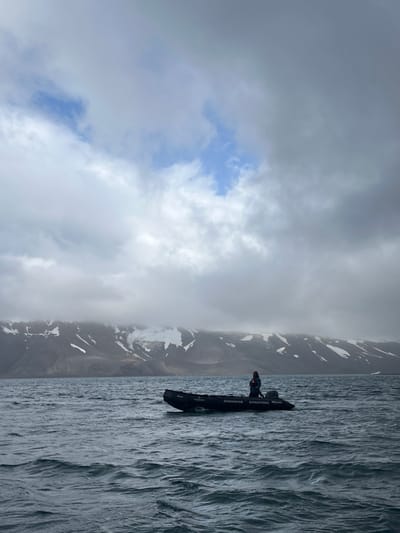 A dramatic landscape with a dark boat on choppy water. A single person sits in the boat, positioned near the shoreline. Mountains rise in the background, partially obscured by low-lying clouds. Above, a mostly overcast sky reveals small patches of blue. The rugged mountain terrain is touched with spots of snow. The image is oriented sideways, with the horizon appearing vertical.