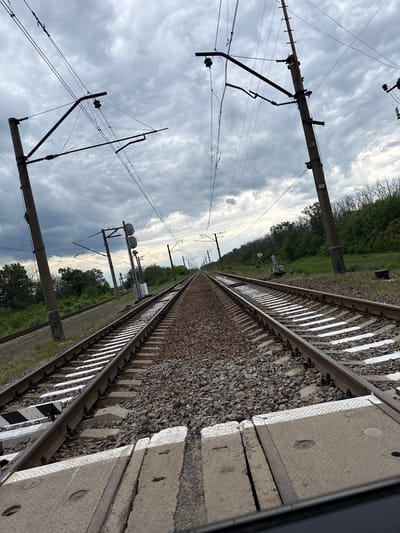 The image displays a straight section of a railway line extending into the distance under an overcast sky with scattered clouds. The perspective is from alongside the tracks, looking forward along their path. At least two parallel tracks are visible, supported by concrete sleepers and surrounded by gravel ballast.
Overhead catenary system poles, constructed from concrete, line the railway, supporting an intricate network of electrical wires, insulators, and support structures. The number "5" is distinctly visible on multiple poles, likely serving as an identification marker. Further along the tracks, several signal posts with multiple signal heads are discernible.
On either side of the railway, green foliage and trees line the horizon, suggesting a rural or semi-rural setting. Near the tracks, a small white box or marker is present. In the foreground, a textured concrete panel, possibly part of a platform or maintenance access, lies adjacent to the rails, featuring circular indentations. A black and white striped marker is also visible on one of the sleepers.
The scene is static, devoid of human activity or trains. It portrays typical railway infrastructure on a bright, cloudy day. The specified location context is Zanky, Ukraine.