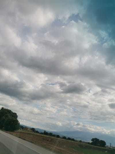 The image depicts a cloudy sky over a rural landscape, likely on the outskirts of Krivina, Bulgaria. The foreground shows a paved road with a white line, angling from the lower left towards the center. Adjacent to the road is a dirt embankment. In the mid-ground, there is a small cluster of buildings with orange roofs, surrounded by trees. Further into the distance, a gentle slope leads to hills and mountains shrouded in a blue haze, under a dramatic sky filled with various shades of gray and white clouds, with a touch of turquoise on the right side. The overall scene conveys a sense of a tranquil, overcast day in a natural setting.