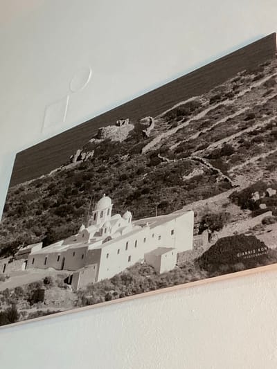 A large, landscape-oriented black and white photograph is mounted on a plain, light-colored wall in Πλατύς Γιαλός, Greece. The photograph depicts a prominent, multi-level white monastery or church complex with multiple domes, situated on a steep, rocky hillside covered with sparse vegetation. Above the structure, terraced dry-stone walls ascend the slope, leading towards remnants of older structures or ruins near the ridge. A body of water, likely the sea, borders the base of the hill to the left. The bottom right corner of the photograph displays faint text reading "GIANNIS KONTOS PHOTOGRAPHY." The wall above the photograph exhibits two faint indentations, one circular and one square, and the photo itself is slightly tilted.