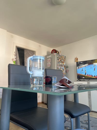 A glass-topped table with two gray chairs is in a room with a window. A water bottle with a white handle sits on the table. A cardboard container is also on the table. A refrigerator with magnets and a television playing a soccer game are in the background. The location is likely in a residence in Benningen, Germany, based on available context. There are no visible people or actions in the frame.