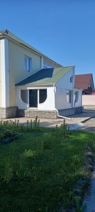 The image shows the exterior of a two-story residential building with a light yellow facade and a green tiled roof. There is a white, enclosed porch or sunroom attached to the front of the building, featuring curved window frames and a pitched roof. The base of the building and the porch are clad in stone. A paved pathway leads to the porch, and a grassy area with some plants is in the foreground. In the background, another building with a brown roof is visible. The sky is clear and blue. The scene depicts a residential property in what appears to be a suburban or rural setting, under a bright, sunny sky. This image is located in the city of Andzhero-Sudzhensk, Russia.