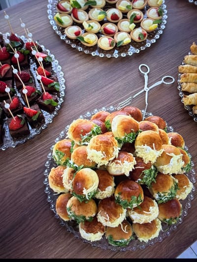 The image captures a meticulously arranged spread of various appetizers and desserts displayed on a dark wooden table, suggesting a catering event or social gathering in Ros Na Rí, Ireland.

In the center, a clear glass platter with a beaded rim is laden with a generous pile of small, round, golden-brown baked buns. Many of these savory items are topped with either shredded white cheese or fresh green herbs, likely parsley or cilantro, offering a blend of textures and flavors.

To the left, another ornate glass plate showcases numerous mini tartlets. These individual pastries feature a creamy filling and are garnished with fresh elements such as bright red strawberry slices, vibrant lemon wedges, and small green mint leaves, adding visual appeal and a fresh zest.

At the bottom of the frame, a third glass platter presents an assortment of square chocolate brownies or cakes. Each piece is elegantly topped with a half strawberry and skewered with a small decorative stick featuring white beads, designed for easy serving.

Partially visible at the top edge of the frame is a fourth glass platter containing a stack of golden-brown rolled pastries, which appear to be either savory rolls or sweet pastry sticks. Resting on the wooden table, positioned between the left and central platters, is a pair of elegant, silver-colored serving tongs with an intricate, decorative design, ready for use.

The overall presentation is neat and inviting, highlighting a variety of small, bite-sized foods suitable for a celebratory occasion. No people are visible, and there is no discernible text within the image. The setting suggests an indoor event, with the time of day being indeterminate due to artificial lighting.
