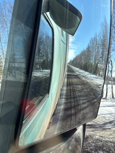 The image presents a roadside view from a moving vehicle in Kukui, Russia. Visible in the left frame is the light teal side panel of a large vehicle, alongside an interior window frame. The prominent right-hand side mirror, soiled with road grime, reflects a receding two-lane road. This road surface is dark, indicating melting snow and dirt, with distinct tire tracks. Flanking the road are snow-covered shoulders transitioning to a dense stand of mostly bare deciduous trees, including apparent birches, with some evergreen specimens interspersed. The sky is bright blue with scattered cirrus clouds, indicative of a clear, sunny day in a late winter or early spring environment. Direct sunlight casts visible shadows. No individuals are depicted.
