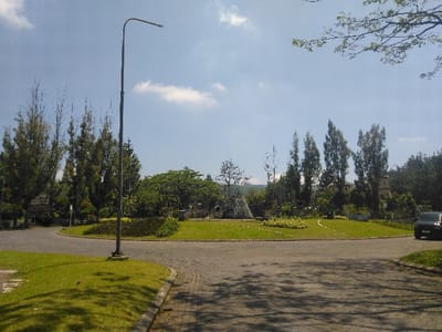 The image depicts an exterior, daytime scene of a traffic roundabout in Nová Bystřice, Czechia, under a clear blue sky with scattered white clouds. A tall, dark gray street light pole with a curved arm and an LED fixture is centrally positioned in the foreground, rooted in a grassy verge. The roundabout features a large, oval-shaped central island, uniformly covered with vibrant green grass. On the left side of this central island, a dark, angular pyramidal structure or monument is visible, partially obscured by surrounding trees. The island is extensively landscaped with various trees, including tall, slender conifers on the left and more robust deciduous trees throughout. Patches of lighter-colored decorative plantings are arranged on the grass towards the right side of the central island. The paved asphalt roadway of the roundabout is visible in the foreground, curving around the central island. On the far right, a dark-colored passenger vehicle, consistent with an SUV or crossover, is partially visible, positioned as if entering or exiting the roundabout. Shadows from an unseen object are cast across the grass in the lower-left foreground. A tree branch extends into the upper right corner of the frame. No people or animals are present. Distant hills are faintly visible on the horizon line in the far background.