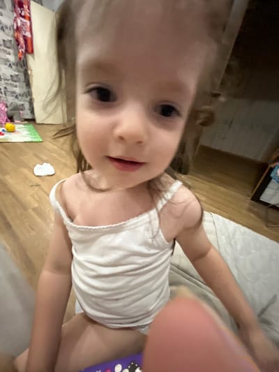 This is a close-up, slightly blurry, selfie-style photo of a young child, likely a toddler, lying down. The child has light-colored hair and is wearing a white tank top. Their face is partially visible, with one eye looking directly at the camera and rosy cheeks. Their mouth is slightly open, as if they are speaking or smiling. The child's arm is extended, and their thumb is prominently in the foreground, obscuring part of the frame.
The background suggests an indoor setting. To the left, there appears to be a wooden floor and part of a shelf or furniture. To the right, a lighter-colored surface, possibly a bed or sofa, is visible. A small, white object, possibly a piece of paper, is on the floor in the lower middle of the image, near a colorful drawing or poster.
The lighting appears to be indoor, possibly artificial, creating some shadows. There is no clear indication of the time of day or weather outside. The overall emotion conveyed is playful and curious, typical of a young child interacting with a camera. No visible text is present in the image. The location context provided is Gorodets, Russia.