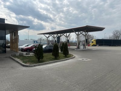 The image depicts an outdoor scene at a commercial service area in Orăștie, Romania, under an overcast sky. The foreground features a grey paved area composed of interlocking pavers, with a landscaped island containing grass and three small evergreen conical bushes separating it from a building on the left.
On the left, a modern building with a dark facade, large glass windows, and vertical wooden slats serving as an architectural feature stands. A white van, a white box truck with "IFL GmbH" lettering, a dark red sedan, and a dark-colored sedan are parked parallel to this building.
In the midground, a large canopy with a red-trimmed roof and a grey metal support structure covers a fueling station. Several white fuel dispensers with dark screens are visible beneath the canopy; no individuals are present at the pumps. To the right of the fueling area, a yellow semi-truck with a black curtain-side trailer is parked or positioned, with an orange traffic cone visible near its cab.
The background shows a line of bare trees on the horizon, suggesting a late winter or early spring season. No individuals are visible engaging in any specific actions within the frame.