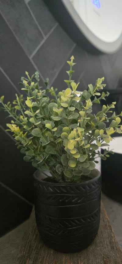 A close-up, eye-level shot shows a small, artificial plant with numerous small, green and yellow leaves. The plant is housed in a dark, cylindrical pot with a textured pattern of horizontal lines and diagonal markings resembling stitches or woven material. The pot sits on a weathered, wooden surface. In the background, a dark gray tiled wall with a herringbone pattern is visible. To the right of the plant, a portion of a white, rounded object, possibly a light fixture or appliance, is present, featuring a faint blue illuminated display. The scene is indoors and appears to be a domestic or commercial setting. The lighting is moderate, with some soft shadows. There are no people or animals depicted. The location is identified as Terrebonne, Canada.