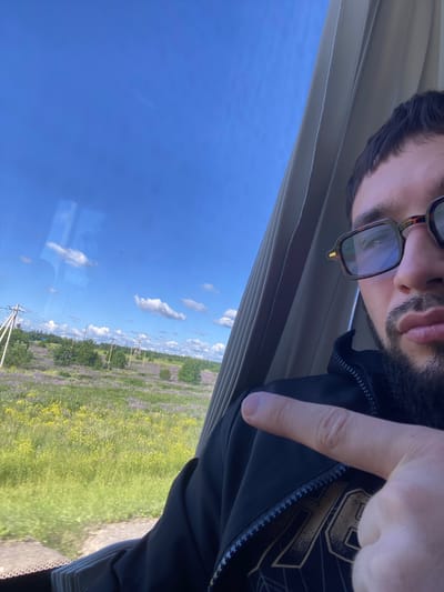 The image captures a selfie taken from the perspective of a person inside a vehicle, likely a bus or train, looking out a window. The foreground is dominated by the right side of the person's face, partially obscured by a finger pointing towards the viewer. The person has short dark hair, a beard, and is wearing glasses with a tortoiseshell pattern. They are also wearing a black jacket.
Through the window, a rural landscape is visible under a bright blue sky with scattered white clouds. The landscape features a field with numerous purple and yellow flowers, interspersed with green grass and trees. Several utility poles and what appear to be power lines run across the field. In the distance, a larger transmission tower is visible. The lighting suggests it is daytime, possibly late morning or early afternoon.
The location is identified as Kipen, Russia. The image conveys a sense of travel and observation of the natural scenery.