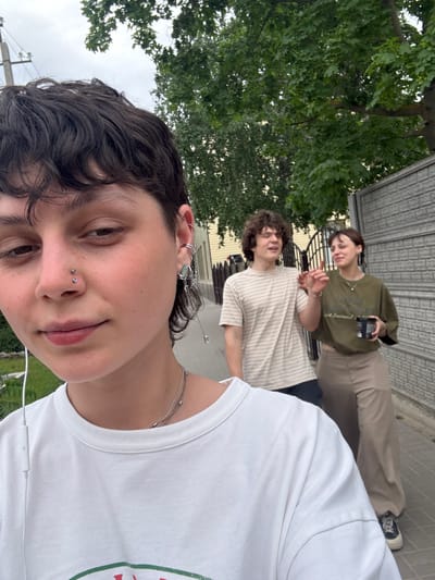 The image is a low-angle selfie. In the foreground, a person with dark, curly hair and a nose piercing looks at the camera, wearing a white t-shirt and a silver necklace. Headphones extend from the person’s ears. Behind them, two other people stand on a paved surface. The person on the left wears a light-colored, horizontally striped t-shirt. The person on the right wears a dark green shirt and khaki pants. Both of them are also looking at the camera. The background features a green tree on the left and a building with metal siding on the right. The scene likely takes place in an outdoor setting. There is no way to determine if this image was taken in Uryupinsk, Russia.