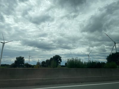 The image depicts a cloudy sky over a rural landscape featuring several wind turbines. In the foreground, a concrete barrier runs horizontally across the frame, with a highway visible in the lower right corner. Behind the barrier, trees and vegetation form a green line, with wind turbines of various sizes dotted across the horizon. The turbines are white with red tips on their blades. The sky is filled with dark, heavy clouds, suggesting overcast weather. The scene appears to be on the outskirts of Großenaspe, Germany, a region known for its wind farms.