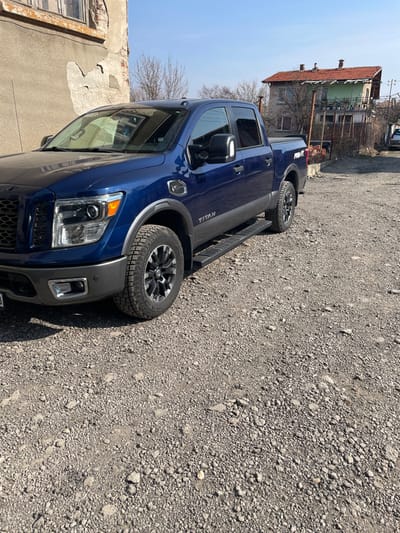 A dark blue Nissan Titan PRO-4X pickup truck is parked on an unpaved gravel surface in Resilovo, Bulgaria, under a clear, bright sky. The vehicle is positioned diagonally, occupying the foreground and midground, with its front bumper and driver's side visible. It features black running boards, black multi-spoke wheels with off-road tires, and front bumper-mounted fog lights/driving lights with integrated circular sensors. "TITAN" badging is present on the lower front fender, and "PRO-4X" is visible on the rear bed side.
To the immediate left and partially obscured by the truck's front, an old, dilapidated two-story building with peeling beige stucco and exposed brickwork stands. Beyond the truck, in the midground and background, bare deciduous trees are visible against the sky. Further in the background, an unpaved track leads towards a two-story residential building with a red tiled roof and a light green upper facade, surrounded by what appears to be a fence and various stored items or debris. Utility poles and overhead lines are present in the distant background. A light-colored vehicle, possibly a white van, is partially visible at the far end of the track. The ground throughout the scene is composed of various sizes of gray and brown gravel. No individuals are present in the image.