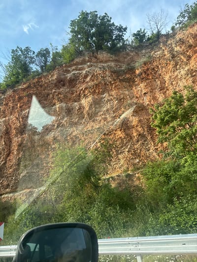 This image is a landscape photograph taken from inside a vehicle in Batovo, Bulgaria, featuring a prominent reddish-brown rock face. The upper section of the image shows a bright blue sky with scattered wispy white clouds. The top edge of the rock face is densely covered with mature green deciduous trees, some with bare branches intertwined.
The main subject is a large, textured rock formation, appearing as an exposed cliff or a geological cut. Its surface exhibits various shades of orange, brown, and lighter tan, suggesting a composition of sandstone or similar rock, with visible striations and erosion patterns. Sparse green vegetation, including grasses and small bushes, adheres to crevices and ledges on the rock face.
In the lower portion of the image, the base of the rock face transitions into a verdant expanse of diverse green foliage, including shrubs, tall grasses, and small white wildflowers.
A large, irregular, bright reflection, likely of the sky or an overhead object, is prominent on the vehicle's window surface, casting a light, diffused area over the mid-left section of the rock face. Additional faint, linear reflections are also visible across the glass.
In the bottom-left corner of the foreground, a portion of a black vehicle side-view mirror is visible, reflecting indistinct shapes. A metallic guardrail extends horizontally across the bottom of the frame, and a fragment of a red and white chevron road sign is partially discernible to the left of the mirror, behind the guardrail. The scene is brightly illuminated by daylight.