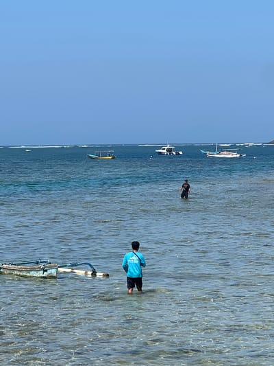 This image depicts a serene coastal scene in Denpasar, Indonesia. The foreground is dominated by shallow, clear turquoise water, revealing the sandy seabed beneath. To the left, a traditional Indonesian outrigger boat, painted in weathered blue and white with blue outrigger poles, rests partially submerged. In the mid-ground, two men are wading in the water. The man closer to the foreground, facing away from the camera, wears a bright turquoise t-shirt and black shorts, with a black strap over his shoulder. The man further away, also facing away, is dressed in a dark t-shirt and dark pants. In the background, several boats are anchored in the ocean. One is a small, yellow and blue traditional boat with a covered seating area. Further out, two modern white motorboats are visible, one with a distinctive hull shape. The horizon is marked by a thin line of white surf and a hazy coastline in the distance under a clear, pale blue sky. The overall atmosphere is tranquil and indicative of a sunny day at the beach.