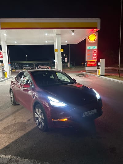A red Tesla Model Y, bearing Bulgarian license plate EA 8899 AB, is parked at a Shell gas station at night in Zagore, Bulgaria. The vehicle is positioned on asphalt pavement, angled slightly towards the right of the frame, with its headlights and front right turn signal illuminated. Overhead lighting from the gas station canopy illuminates the scene. A prominent Shell sign with a glowing yellow shell logo is visible, displaying fuel prices: FuelSave 95 at 1.59, FuelSave Diesel at 1.68, and AutoGas at 0.75. Gas pumps are visible on the left side of the frame. In the dark background, other vehicles are discernible behind a barrier, illuminated by distant lights. No individuals are visible in the image.