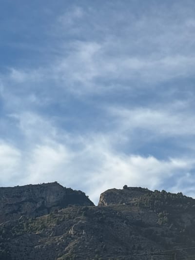 The image depicts a mountainous landscape under a blue sky with scattered white clouds. Two prominent mountain ridges occupy the lower half of the frame, separated by a distinct geological depression. The ridge on the left displays rugged, dark rock formations with sparse vegetation. The ridge on the right features exposed rock leading to a peak, with its slopes and summit densely covered in coniferous trees. The sky above is light blue, traversed by elongated, wispy white clouds. A faint, dark, horizontal line is present at the very bottom edge of the frame, indistinct in detail. No people, animals, or clear man-made structures are visible. The terrain is consistent with the mountainous region surrounding Alcoi/Alcoy, Spain.