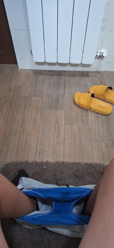 This is a medium shot, taken from a high angle, of a bathroom floor. In the top left corner, a white radiator is visible, attached to a tiled wall. On the wooden tiled floor, a pair of yellow slide sandals are placed. The right side of the image shows a person's legs with blue underwear pulled down, resting on a brown, shaggy rug. The scene is likely indoors in a residential setting in Studena, Bulgaria, and the lighting suggests it is daytime. There are no discernible emotions or weather indicators. The text "Cupra" and "Summe" are visible on the yellow sandals.