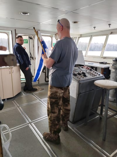 The image appears to be taken inside the bridge of a boat. The scene includes two men; one is holding a flag with a blue and white striped design. The other man is leaning against a cabinet. The flag-holder is wearing a grey t-shirt and camouflage pants. The man leaning against the cabinet is wearing a dark blue jacket. The bridge has a control panel, a stool, windows showing a landscape with bare trees, and a sign above a doorway that says "EXIT". Based on the available information, it is not possible to confirm that this image was taken in Vskhody, Russia.
