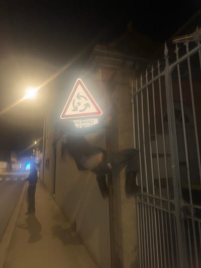 The scene captures a nighttime urban environment in Roanne, France, under artificial illumination. In the central foreground, a reflective white triangular road sign with a red border displays a black symbol of three arrows circulating a central dot, indicative of a roundabout. Directly below this sign, a white rectangular sticker with black text, partially obscured by light, reads "VOUS N'AVEZ LA PRIORIT?" (You do not have the priority).

To the right of the sign, an individual is in the process of scaling a light-colored stone pillar or wall, which stands adjacent to a dark metal gate. This individual wears dark outerwear and dark trousers; a portion of light-colored undergarment and skin is visible on their lower back, exposed above the waistline of their trousers. Their right leg is bent, with a dark boot resting on one of the horizontal bars of the metal gate. The individual's upper torso extends over the top of the pillar.

Further down the street on the left side of the image, a second individual stands upright on a paved sidewalk. This person is dressed in dark clothing and is positioned with their back or side oriented towards the camera.

The environment includes a bright street lamp on the far left, generating a significant lens flare that largely obscures portions of the left side of the image and the road sign. The paved sidewalk extends into the background, bordering a building with a visible facade and cornice. A street with white lane markings is partially visible beyond the sidewalk. The metal gate features multiple vertical bars and pointed finials along its top edge, serving as a barrier to a property.