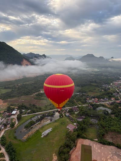 An aerial, wide-angle shot captures a vibrant red and yellow hot air balloon ascending over a misty, mountainous landscape at dawn. The balloon is in the foreground, dominating the right side of the frame. In the mid-ground, a village with scattered houses and green fields is visible, bisected by a narrow road. A winding river or canal cuts through the landscape, lined with trees and small buildings. The background features lush green mountains shrouded in a layer of mist, with darker, more distant peaks visible on the horizon. The sky is overcast with diffused sunlight breaking through the clouds. The scene appears to be Hong Ngern Village, Laos.