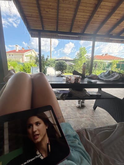 The image depicts a person's legs and a tablet screen on a patio, likely in Viden, Bulgaria. The setting is outdoors, under a wooden-covered patio with a view of a backyard and houses. The time of day appears to be sunny, with a bright blue sky filled with white clouds. The patio itself has a tiled floor and is furnished with dark wicker chairs and a table. On the table, there are a few objects, including a bottle, a container of ice cream, and a towel. A light string of fairy lights is draped across the patio ceiling.
The main focus of the image, besides the person's legs, is the tablet screen which displays a video or image of a woman. The woman on the screen has dark hair and is looking towards the camera with a serious expression. Visible text on the tablet screen appears to be in Cyrillic script. The person holding the tablet is partially visible, with their legs positioned in a relaxed manner, suggesting they are sitting or lounging. A light blue towel is draped over their legs. The overall mood is casual and relaxed, suggesting a leisure activity on a pleasant day.