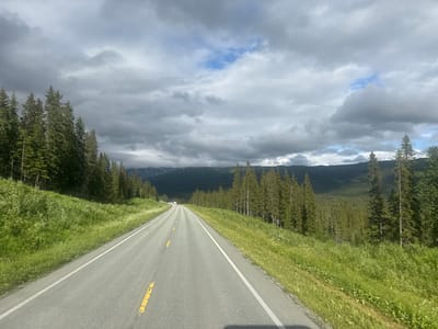 This media file shows a scenic drive through a mountainous, forested landscape. The perspective is from a vehicle moving along an asphalt road that curves gently into the distance. The road is marked with a solid white line on the right edge and broken yellow lines on the left, indicating it's a two-lane highway.
On either side of the road, lush green grass and wildflowers cover the slopes, interspersed with tall, dark green pine trees. The dense forest extends up the sides of the mountains, which form the backdrop of the scene. The mountains themselves are covered in trees and appear to have snow-capped peaks in the distance, suggesting a high-altitude or northern location.
The sky is dramatic, filled with a mixture of dark, heavy clouds and patches of bright blue sky, indicating changeable weather, possibly an overcast day with some sun breaks. The lighting suggests it is daytime, likely afternoon, due to the direction of shadows and the overall brightness.
In the distance, a few vehicles can be seen on the road, including what appear to be a couple of white vans or campers, and possibly a smaller car. There are no people clearly visible in the image. The overall mood conveyed is one of peaceful travel and immersion in nature.
The location is identified as Storforshei, Norway, which aligns with the typical landscape of mountainous regions in Norway characterized by forests, roads, and dramatic skies. No text is visible in the image.