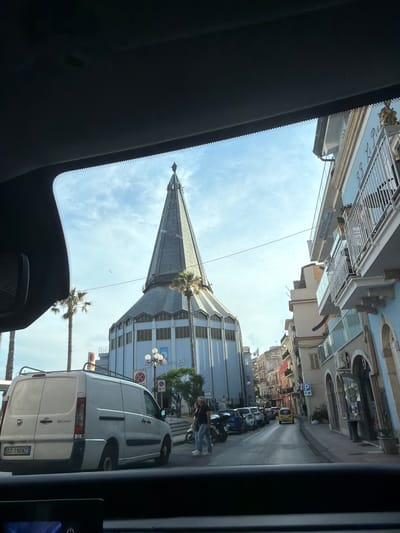 A view from inside a vehicle shows the Basilica of St. Peter and Paul towering above the street in Chianchitta, Italy. The basilica's gray spire dominates the skyline. The street scene includes parked motorcycles and a white Fiat van. Pedestrians are present near the vehicles. Buildings line the street on either side. The sky is light blue with scattered clouds. The vehicle's interior elements are visible in the foreground.