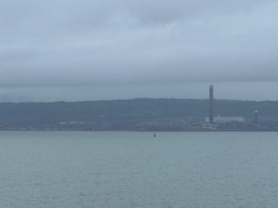 The image depicts a panoramic view across a body of water towards a distant landmass and industrial complex, visible from or within the city of Bangor, United Kingdom (specifically, Bangor, Northern Ireland). The scene is dominated by a uniformly overcast, light grey sky in the upper third, transitioning to a calm, grey-blue body of water occupying the lower half of the frame, showing minor surface ripples.
A red, conical marker buoy is present in the foreground water, positioned slightly to the left of the image's vertical center. Beyond the water, a dark, low-lying landmass stretches horizontally across the middle ground, appearing hilly or undulating with indistinct patterns of vegetation and sparse urban development.
Prominently located on this distant landmass, slightly right of center, is a large industrial power generation facility. The facility comprises a very tall, slender, grey cylindrical chimney or stack, which rises vertically from a cluster of lower, light-colored rectangular industrial buildings. A second, shorter, and thinner vertical stack is visible to the right of the main complex. No smoke, emissions, or active machinery are discernible from the facility. No people or animals are visible in the scene.