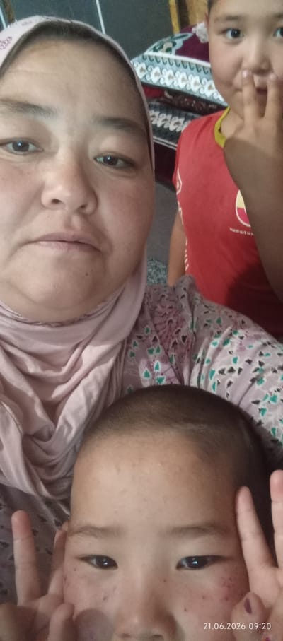 A close-up selfie taken in Chon-Kara, Kyrgyzstan, shows three individuals. The primary subject, in the foreground, is a young child with short dark hair, facing the camera. To the left and slightly behind is an adult woman wearing a pink hijab and a patterned top, her face partially visible. To the right and further back, another child in a red shirt is visible from the chest up, with one hand raised towards their face. The background appears to be an indoor setting with what looks like patterned blankets or textiles. The timestamp "21.06.2026 09:20" is overlaid on the bottom right of the image.